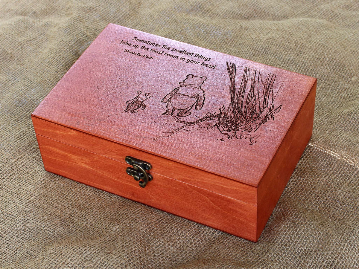 A rectangular wooden keepsake box in mahogany finish, featuring an engraved Winnie the Pooh illustration and quote on the lid, resting on a textured beige fabric background.